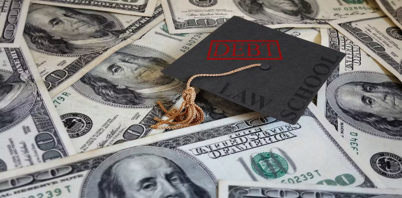 $100 dollar bills scattered around with a graduation cap laying on top that reads, "DEBT."